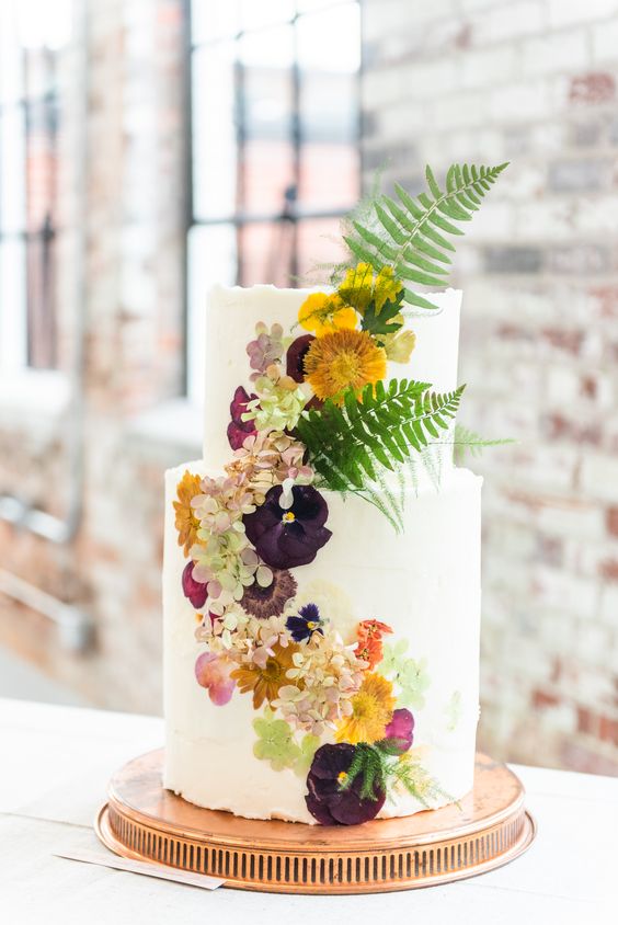 a white buttercream wedding cake with uneven edges and bold mustard, pink and deep purple blooms pressed and some leaves standing out for a fall wedding