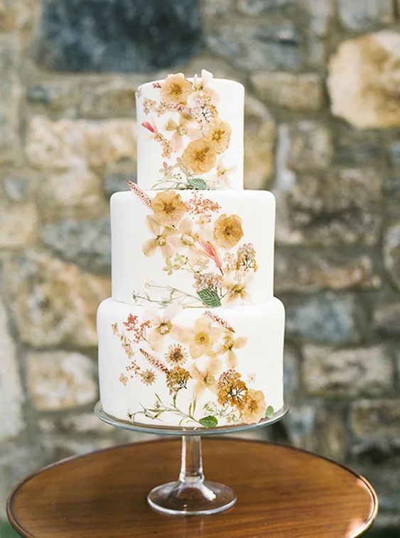a breezy white buttercream wedding cake with pastel yellow and blush flowers and greenery covering the front of the wedding cake