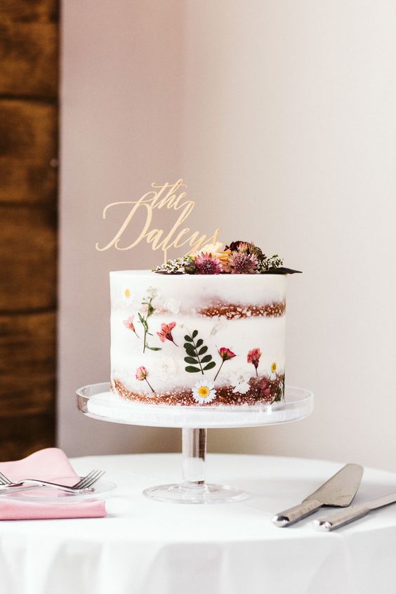 a gorgeous naked wedding cake with a bit of white and pink blooms and a leaf plus dried flowers on top and a calligraphy topper
