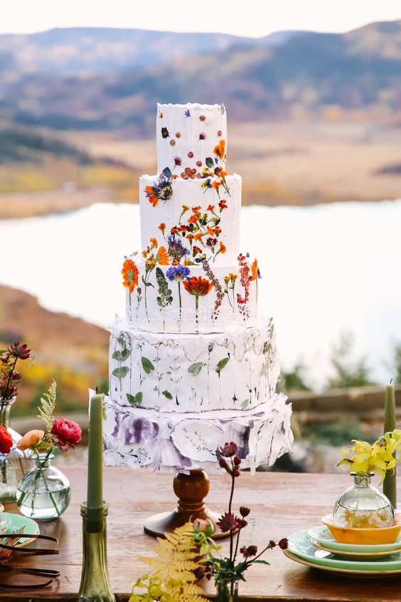 a multi tier white wedding cake with pressed bold blooms, a tier with only leaves and a painted lilac tier is amazing for summer