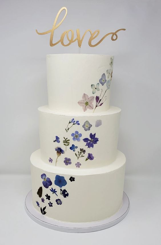 a white buttercream wedding cake with a delicate pattern o blue and purple blooms going up and a calligraphy topper