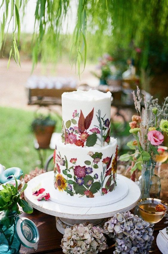 a white buttercream wedding cake with uneven edges and mustard, red and pink blooms and leaves pressed to it to form a pattern