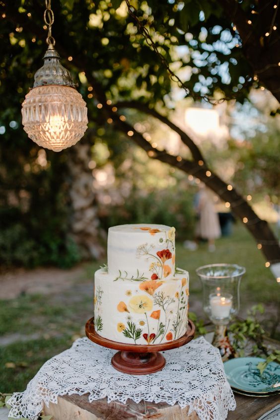 a bright and cool white buttercream wedding cake with orange and red pressed flowers and greenery for a summer boho wedding