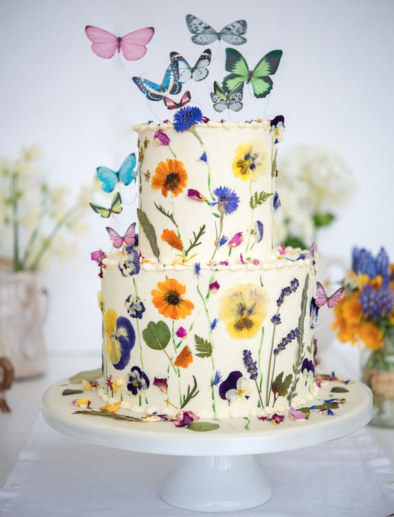 a fantastic white buttercream wedding cake with uneven edges, colorful pressed flowers and leaves and colorful butterflies as cake toppers