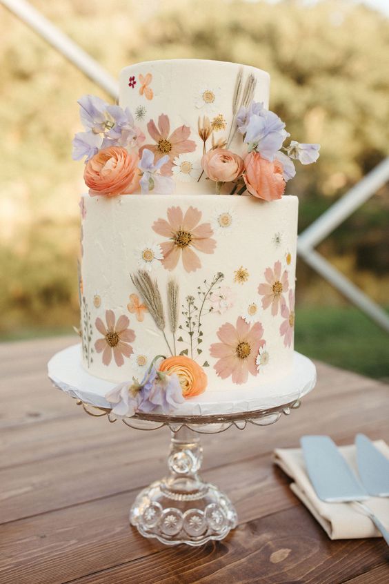 a lovely white buttercream wedding cake with wihte and blush pressed flowers and lilac and orange blooms on top for a spring celebration