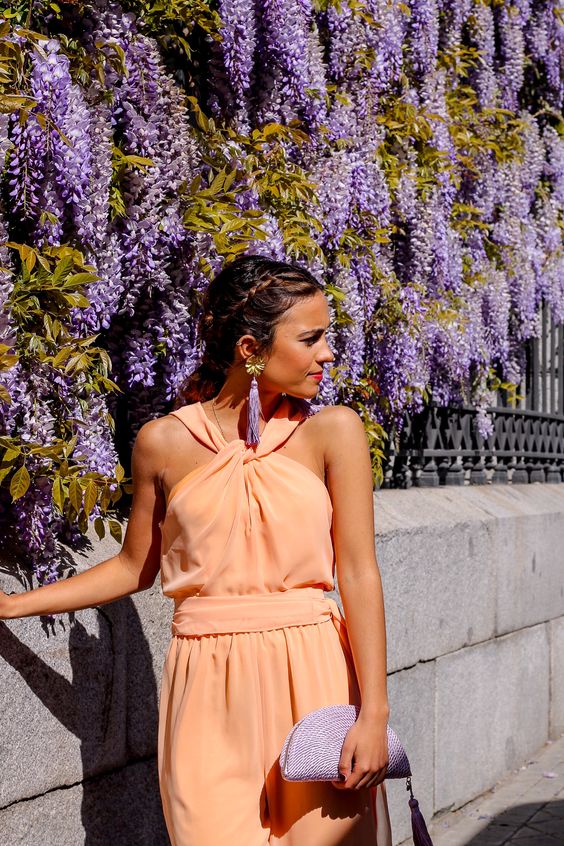 a soft orange jumpsuit with a catchy neckline, lilac tassel earrings and a lilac clutch with a tassel for a spring or summer garden wedding