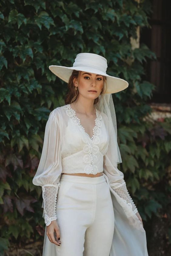 a unique bridal outfit with a vintage inspired lace crop top with puff sleeves and high waisted pants, a white hat with a veil is amazing