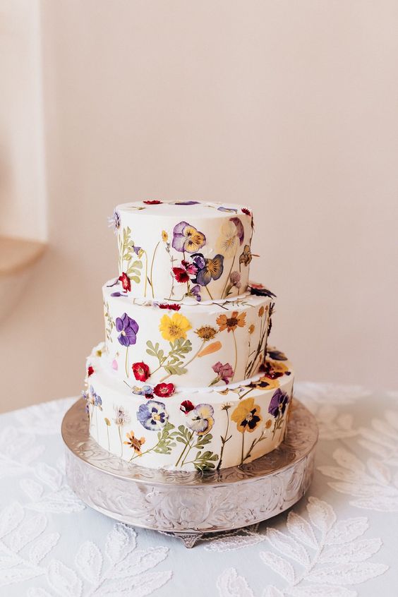 a white buttercream wedding cake with colorful pressed flowers and leaves is a cool idea for a bright boho wedding in summer