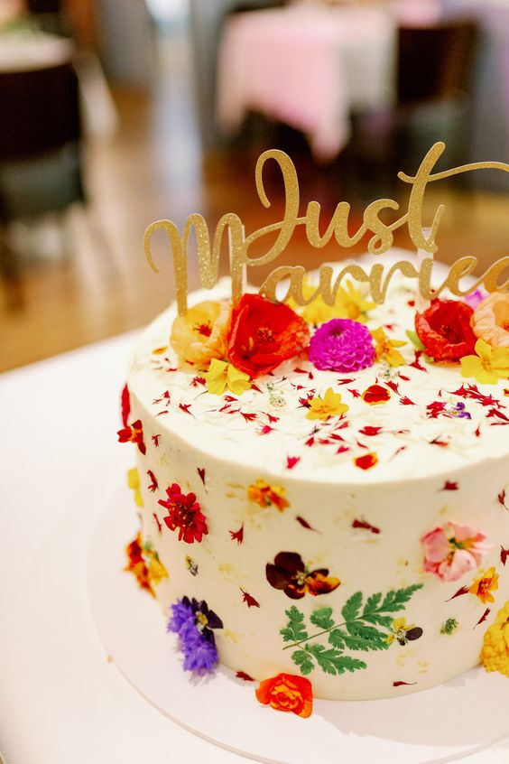 a white buttercream wedding cake with colorful pressed flowers and leaves, with bold blooms on top and a gold glitter calligraphy topper