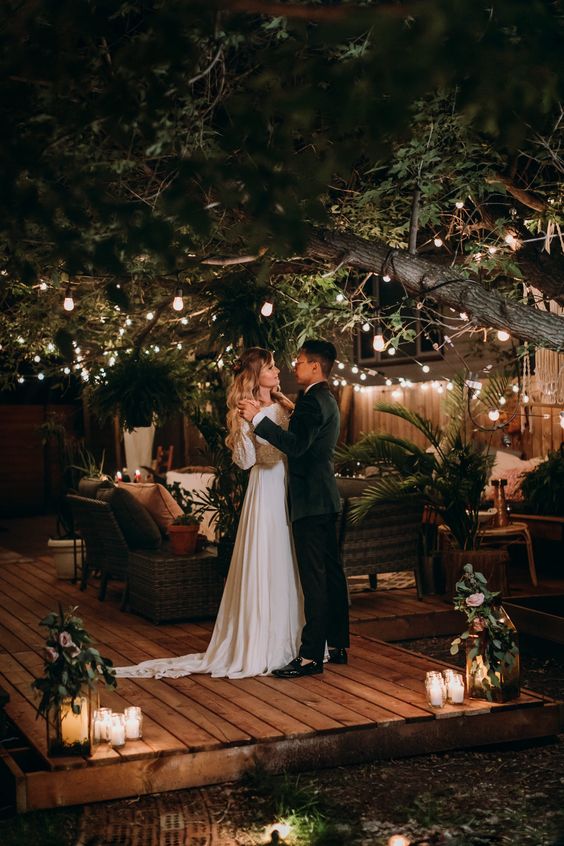 an evening wedding lounge with lights hanging on the trees, candle lanterns on the floor and potted plants and greenery