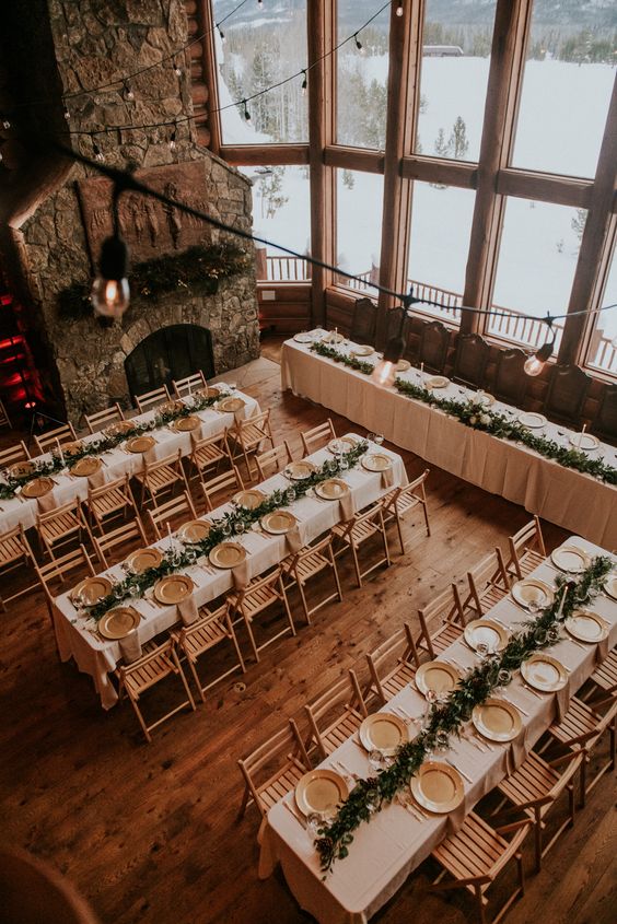 a beautiful chalet wedding reception with a stone fireplace, textural greenery runners, gold chargers and lights over the space