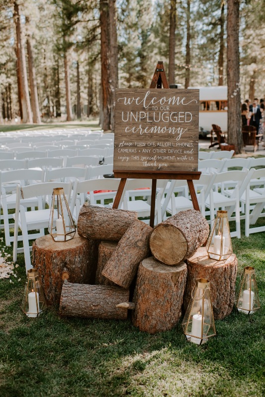 a cozy and lovely wedding ceremony space with white chairs, petals on the ground, tree stumps, candle lanterns and a sign