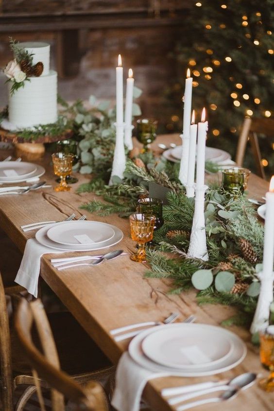 a gorgeous chalet wedding table setting with an evergreen and pinecone table runner, tall candles, white porcelain and amber glasses