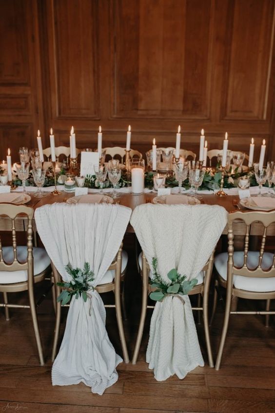 a refined chalet wedding tablescape with a white and greenery runner, candles in gilded candelholders, pillar candles and blankets on the chairs