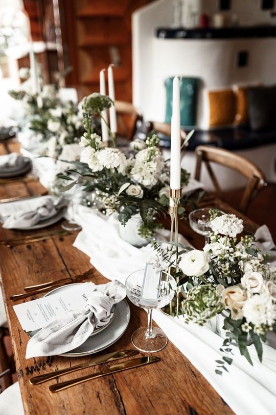 a rustic chalet wedding tablescape with neutral linens, lush white florals, tall and thin candles, white porcelain and gold cutlery