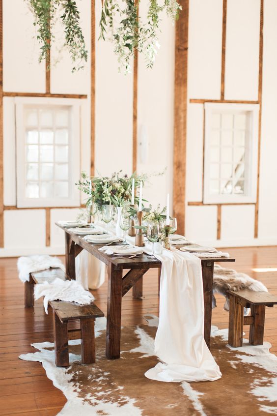 a simple and cozy chalet wedding reception space with neutral linens, greenery, tall and thin candles and benches with faux fur
