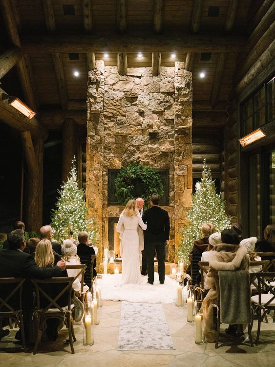 a winter chalet wedding ceremony space with a stone fireplace, Christmas trees, an evergreen wreath with pinecones and candles