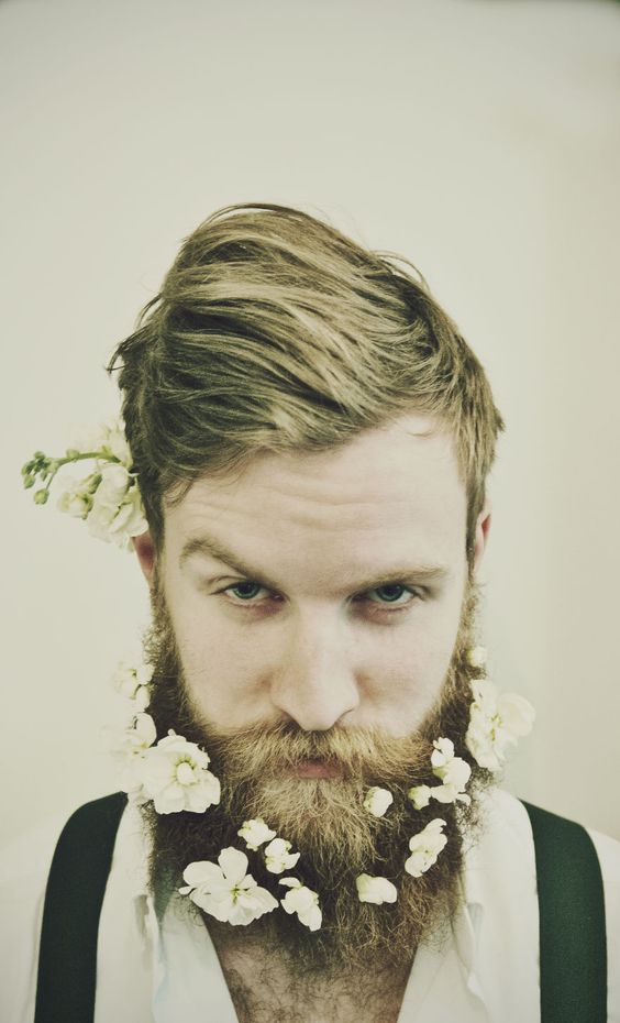 a hipster groom wearing a white shirt and black suspenders plus a beard tucked with white blooms looks unusual