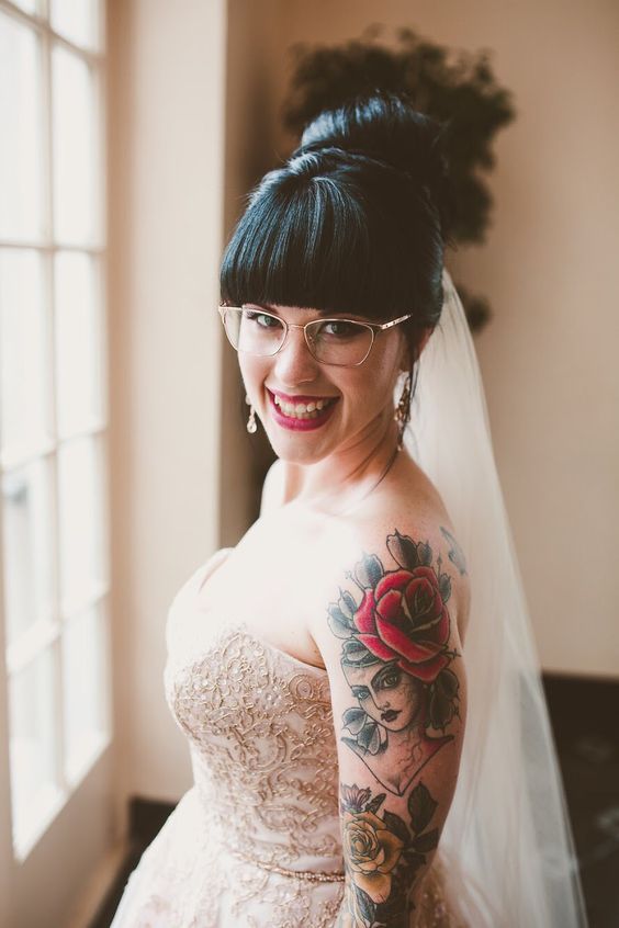 a romantic bridal look with a blush strapless wedding dress, bold bride's tattoos shown off, delicate nude glasses and chic earrings