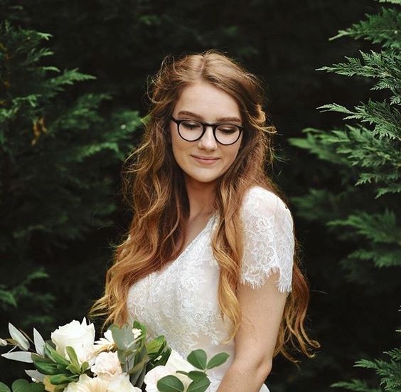 a romantic bridal look with a lace top with short sleeves, a wavy half updo and black round glasses