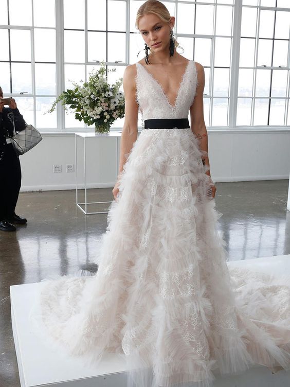 a romantic lace A-line wedding dress with a plunging neckline, no sleeves, a black sash for an accent and black earrings by Marchesa