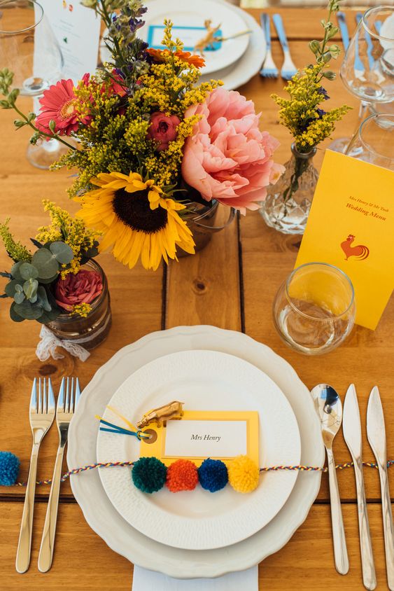a bright and relaxed summer wedding tablescape with a bold card and pompoms for an accent and a pink and yellow flower centerpiece