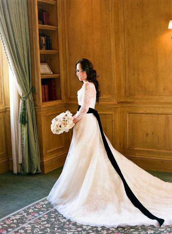 a classic and refined white lace wedding dress with illusion sleeves and a train, a long black ribbon sash and a cutout back with buttons