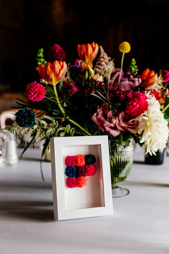 a bright wedding centerpiece with orange, pink, fuchsia and white blooms and greenery, with a colorful pompom applique for decor