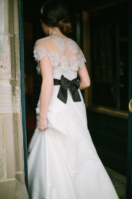 a romantic bridal look with a white strapless A-line wedding dress, a lace capelet over it and a black ribbon bow on the back