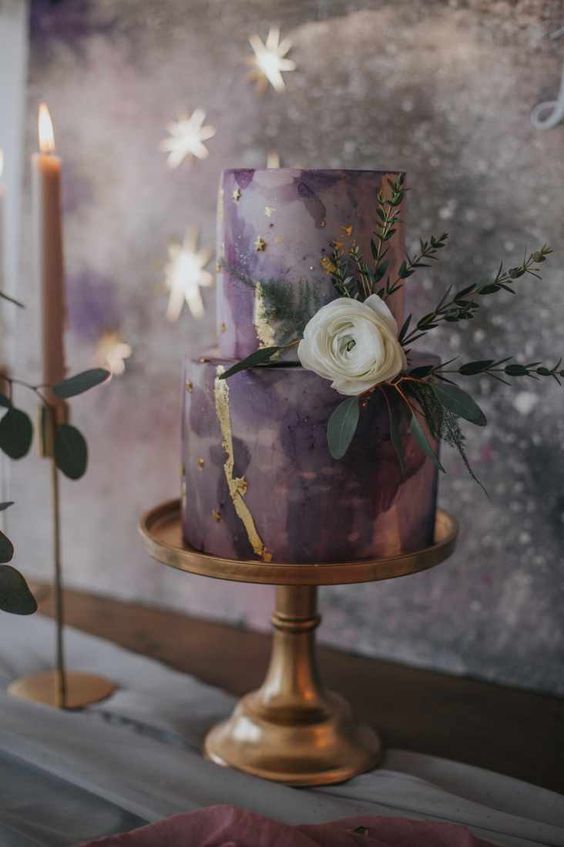 a dreamy celestial wedding cake done in the shades of purple and lilac, with gold glitter and foil, with a white bloom and greenery