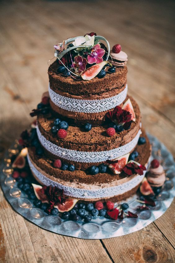 a naked boho wedding cake topped with fresh berries and figs, with blooms and greenery on top plus white lace ribbon decor