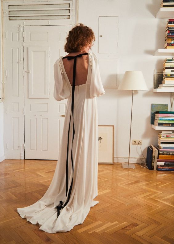 a dreamy flowy white wedding dress with bell sleeves, a pleated skirt, an open back with a black ribbon and a bow to accent it
