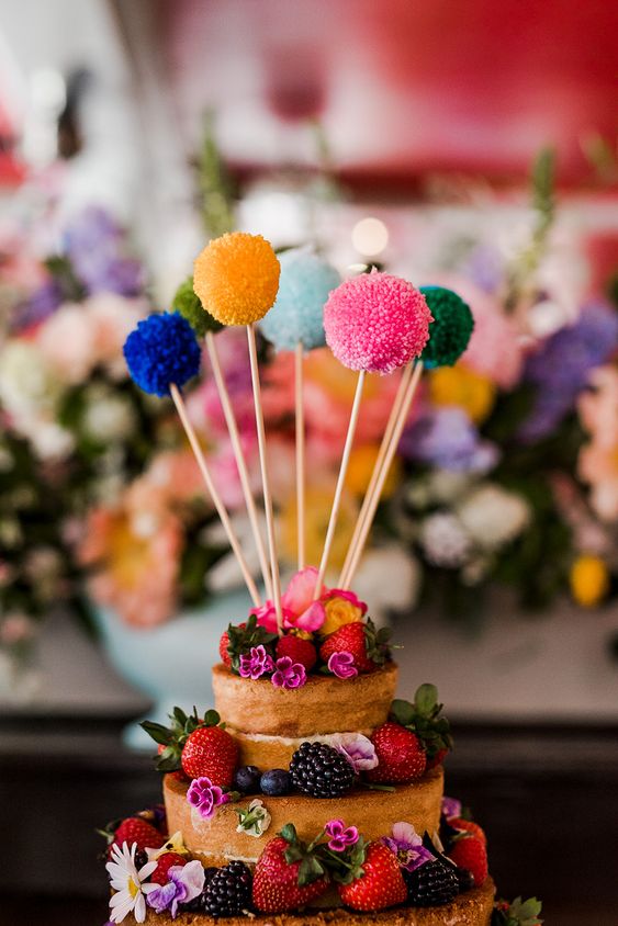 a naked wedding cake with berries and blooms, with colorful pompoms on sticks is a lovely idea for a cheerful party wedding