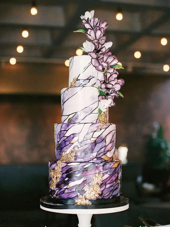 a jaw-dropping wedding cake with purple and black patterns painted on it, with gold foil and gorgeous purple blooms on top