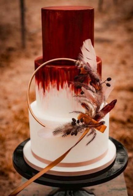 a boho fall wedding cake with a red and white tier, with textures, a round wreath with feathers and berries is amazing
