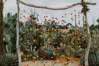 20 a super colorful wedding arch of branches, with greenery, colorful pompoms and tassels on the ends, with a bright striped rug