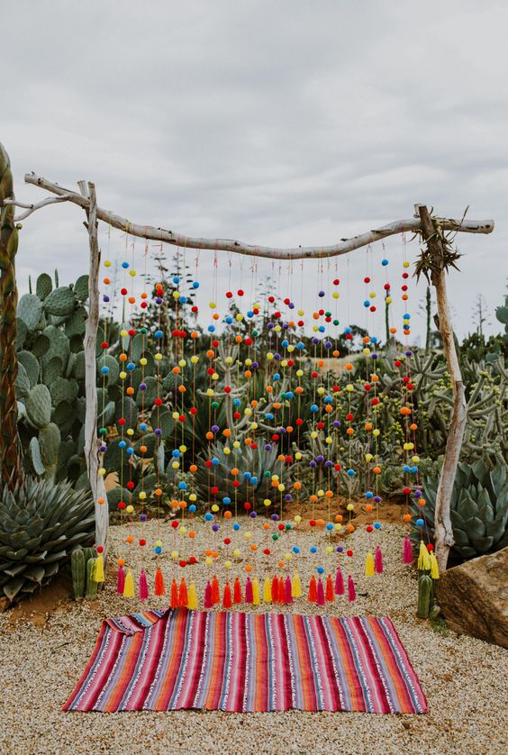 a super colorful wedding arch of branches, with greenery, colorful pompoms and tassels on the ends, with a bright striped rug