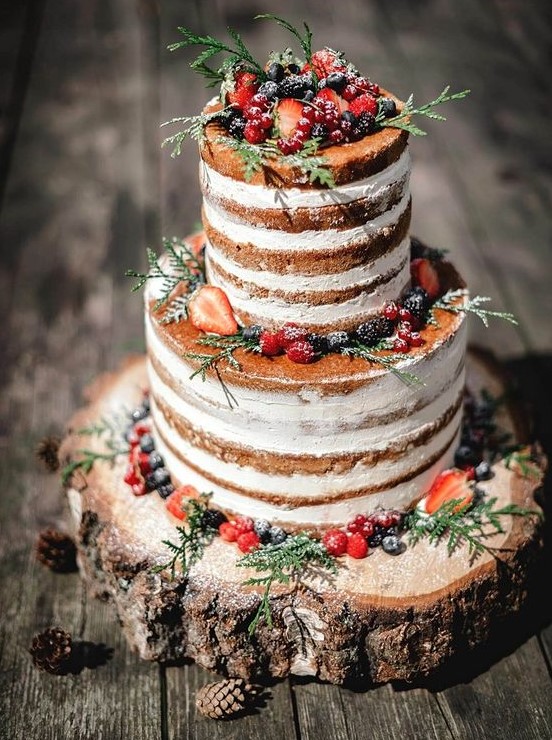 a naked wedding cake topped with sugared berries, greenery served on a wood slice for a rustic wedding