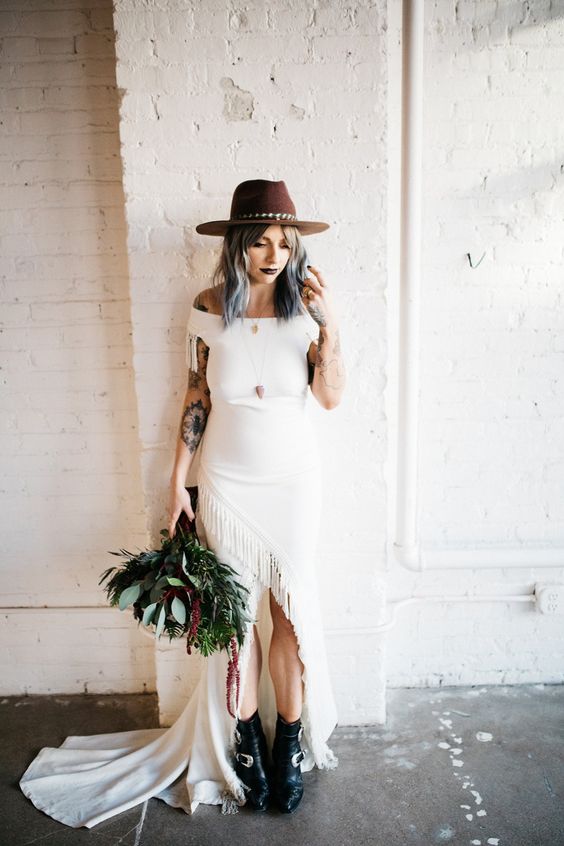 a plain off the shoulder boho wedding dress with fringe, a train, a brown hat, black boots with buckles and a greenery bouquet