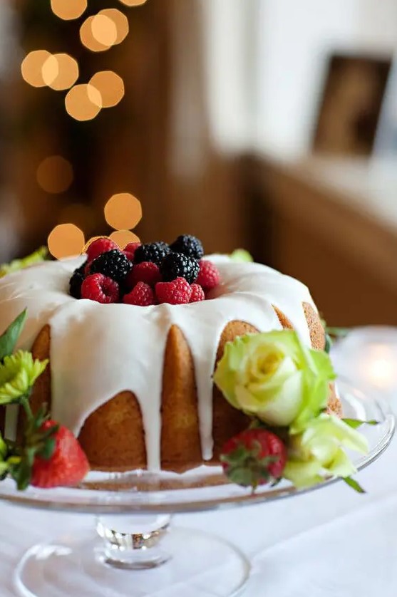a bundt wedding cake with creamy drip, fresh berries on top and yellow roses and strawberries