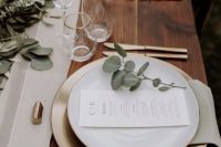 a beautiful and delicate modern wedding tablescape with a neutral table runner and lots of eucalyptus, with gold chargers and cutlery with white menus