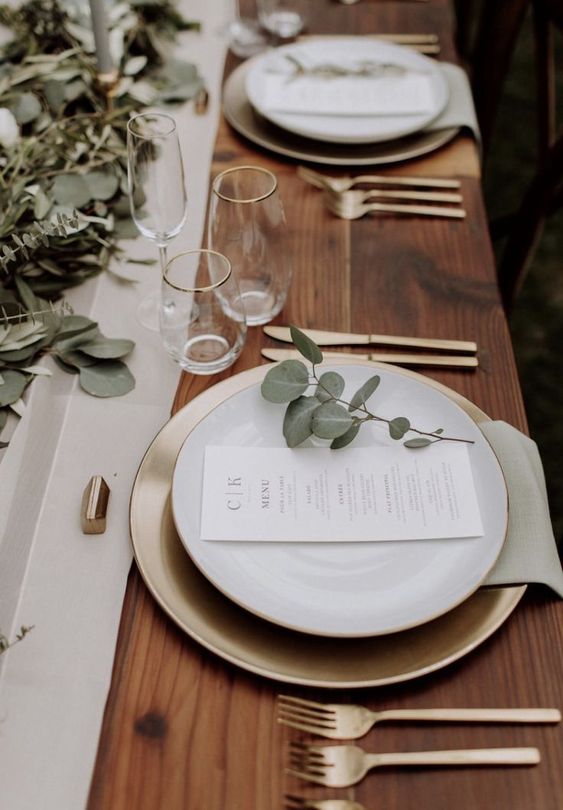 a beautiful and delicate modern wedding tablescape with a neutral table runner and lots of eucalyptus, with gold chargers and cutlery with white menus