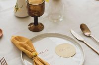 a beautiful modern wedding tablescape with a neutral tablecloth, a mustard napkin, a yellow and mustard centerpiece and a brown glass