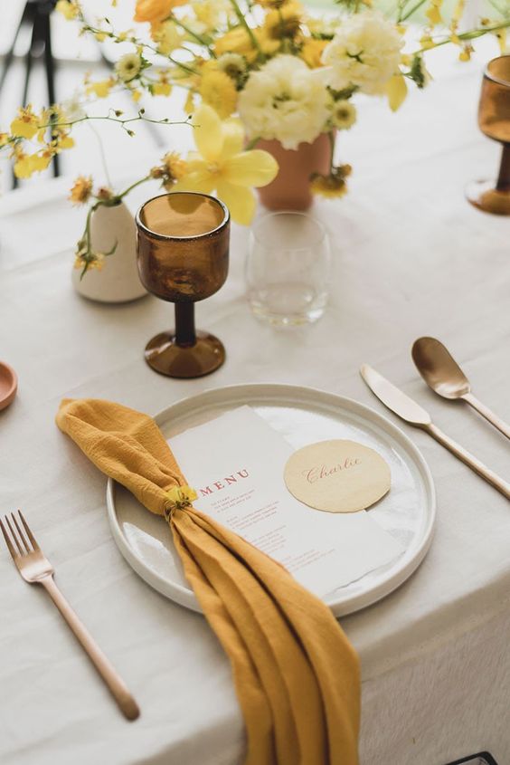 a beautiful modern wedding tablescape with a neutral tablecloth, a mustard napkin, a yellow and mustard centerpiece and a brown glass