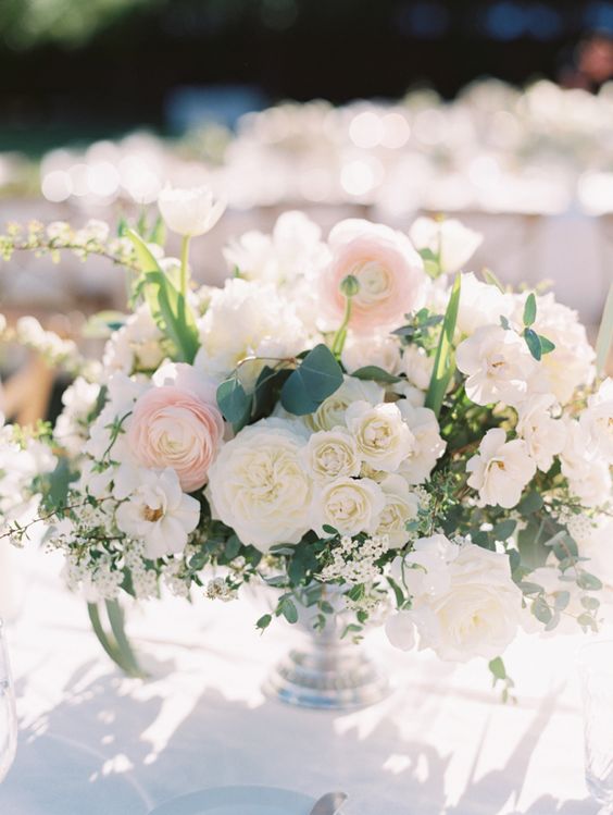 a beautiful wedding centerpiece of neutral and blush blooms, wiht roses and ranunculus, greenery in a silver bowl