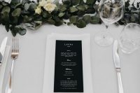 a chic modern wedding table setting with a greenery and white bloom runner, neutral linens, black menus and silver cutlery is cool