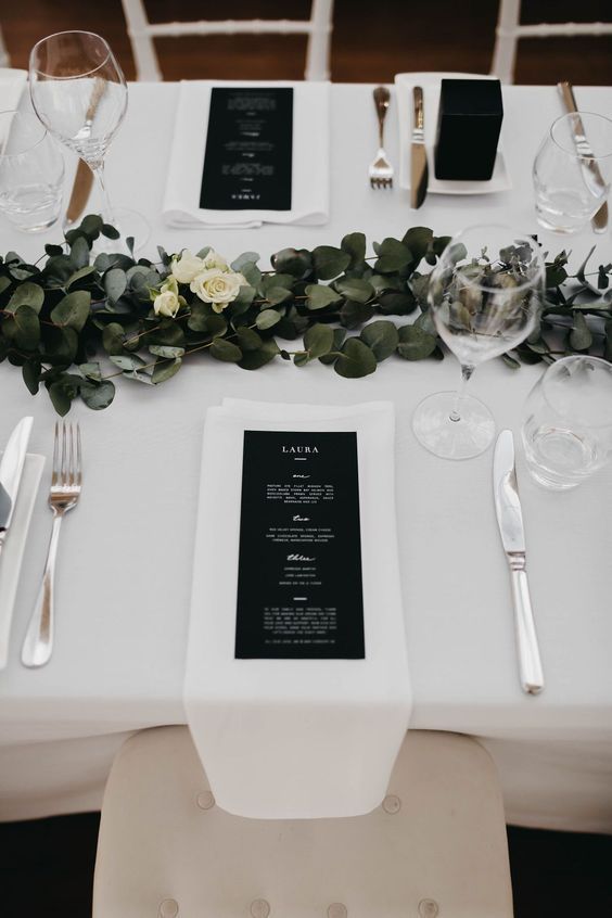 a chic modern wedding table setting with a greenery and white bloom runner, neutral linens, black menus and silver cutlery is cool