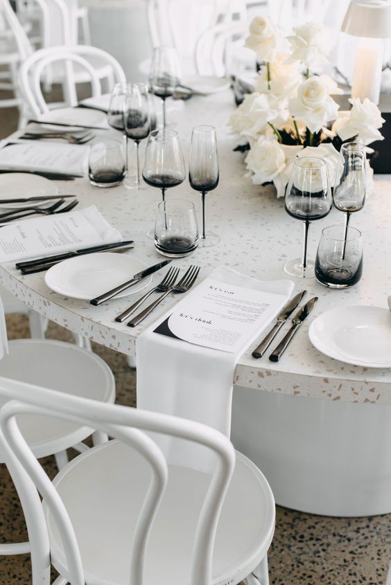 a chic modern wedding tablescape with a terrazzo table uncovered, white napkins, large menus, white roses and black glasses