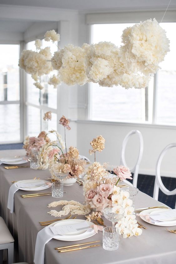 a chic neutral wedding tablescape with a grey tablecloth and white napkins, white hydrangeas, orchids and blush roses and gold cutlery