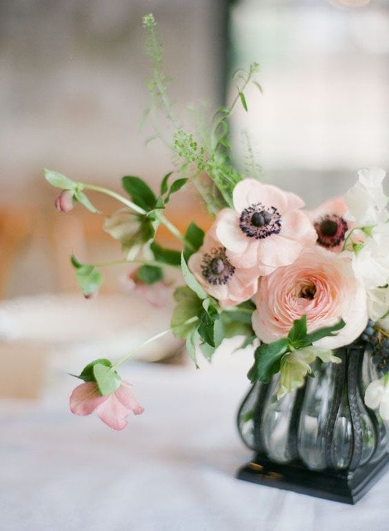a chic wedding centerpiece of a glass vase with black framing, peachy pink ranunculus and anemones, greenery and mauve blooms is wow